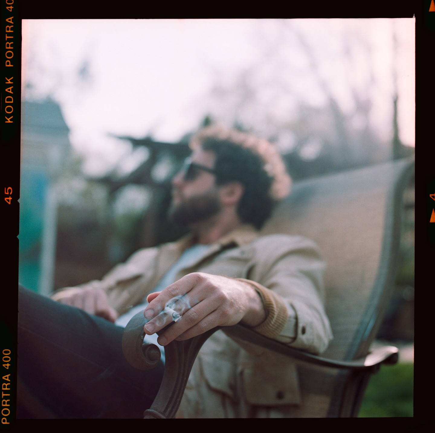 A man with curly hair and sunglasses sits in a chair, holding a lit cigarette.