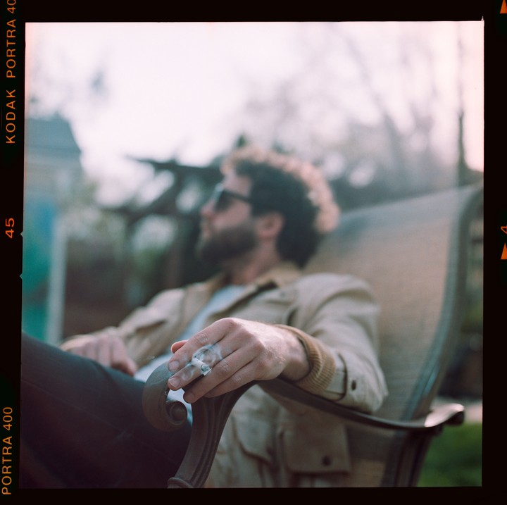 A man with curly hair and sunglasses sits in a chair, holding a lit cigarette.