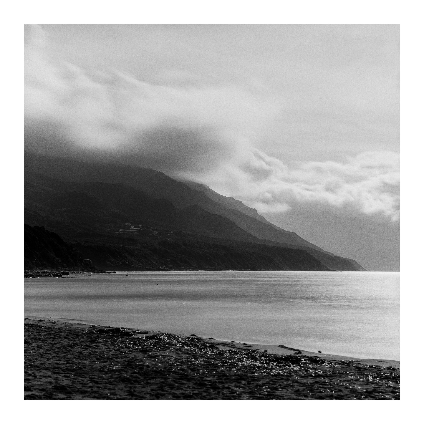 Clouds hang over mountains that meet a calm body of water and a sandy beach.