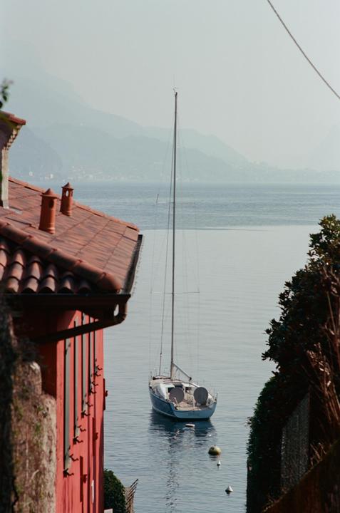 A sailboat floats on a lake with mountains in the background, framed by a red building and foliage.