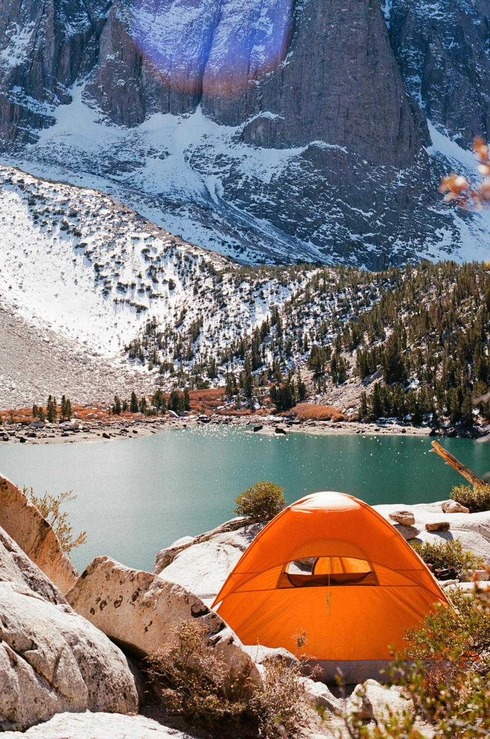 An orange tent is pitched on a rocky shore next to a lake with snow-covered mountains in the background.