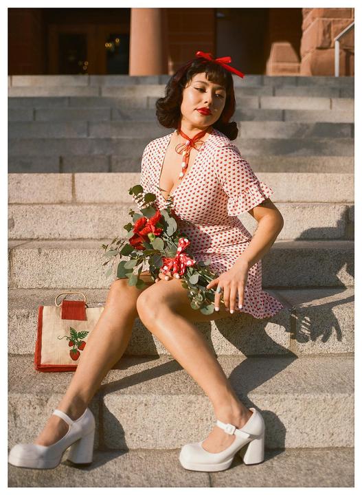 A woman in a heart-patterned dress sits on stone steps, holding a bouquet of red flowers and eucalyptus.