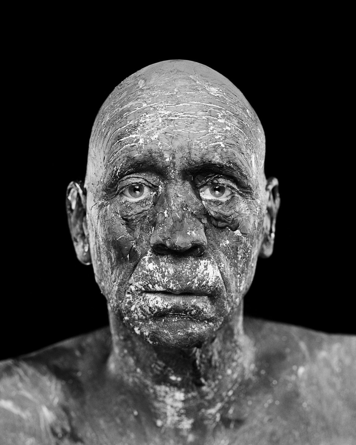 A man with a bald head and face covered in cracked clay looks directly at the viewer.
