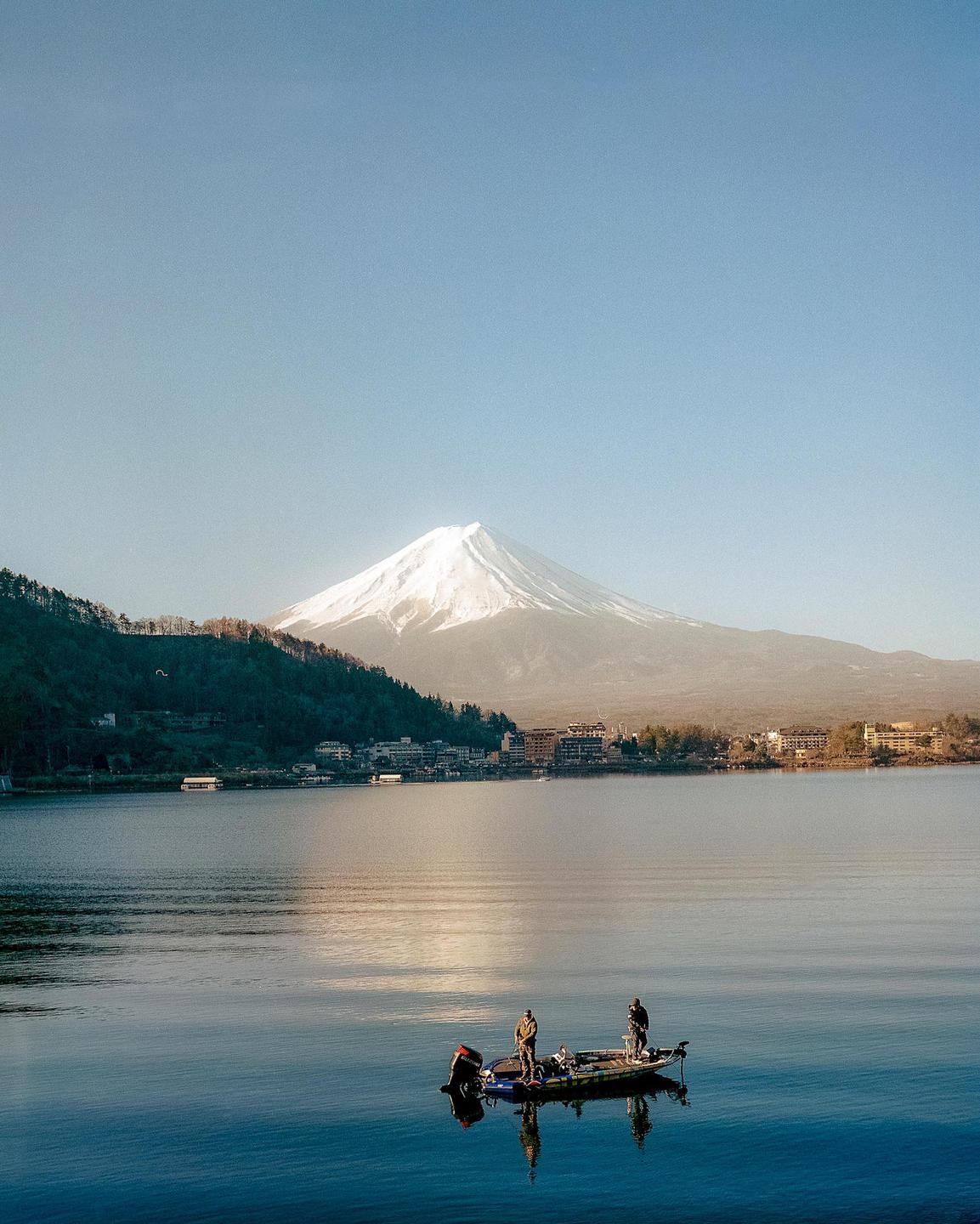 Two men stand in a boat on a lake with a snow-capped mountain and a town in the background.