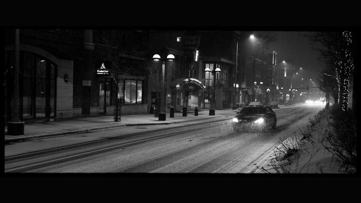 A car with its headlights on drives down a snowy street at night, with buildings and streetlights visible on either side.