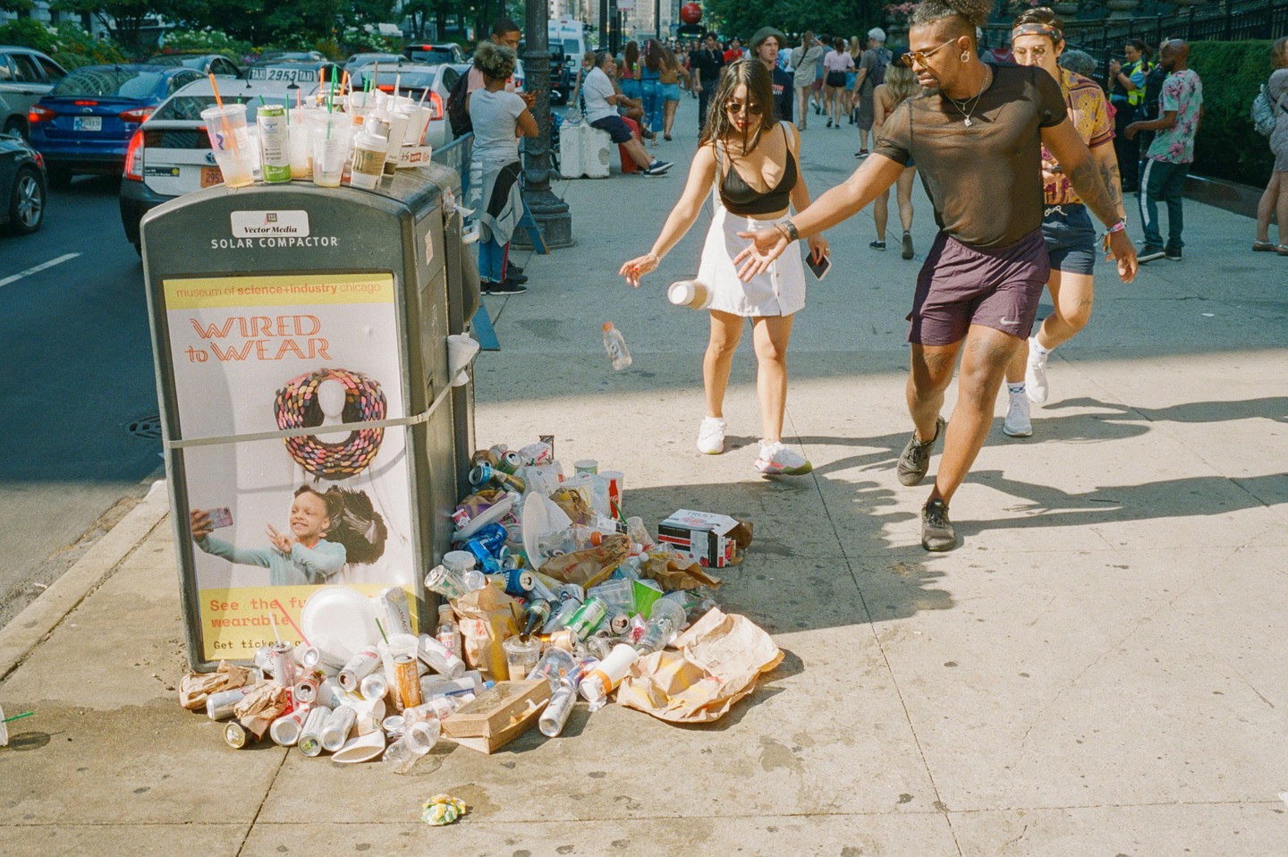 A woman drops a bottle next to an overflowing trash can on a city street.