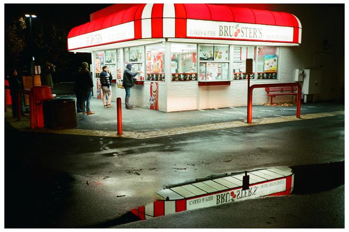 A group of people stand outside an ice cream shop at night, with the shop's reflection visible in a puddle.