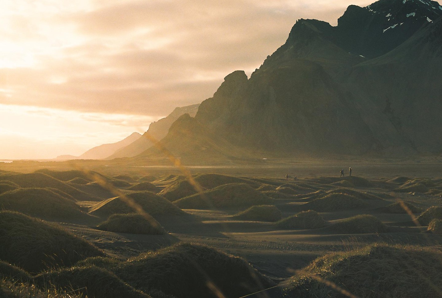 Sunlight illuminates a vast landscape of sand dunes and mountains with a few people in the distance.