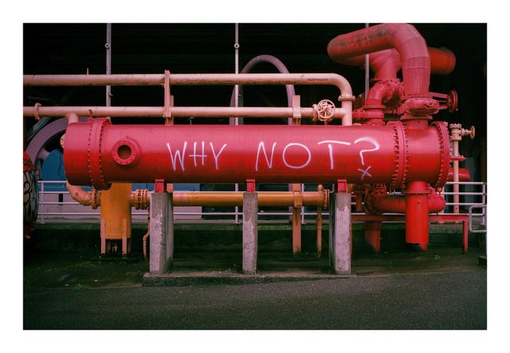 A large red pipe with "WHY NOT?" written on it in white is surrounded by other pipes and industrial structures.