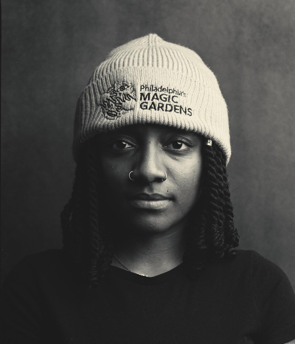 A woman with dreadlocks and nose rings wears a beanie with "Philadelphia's Magic Gardens" written on it.