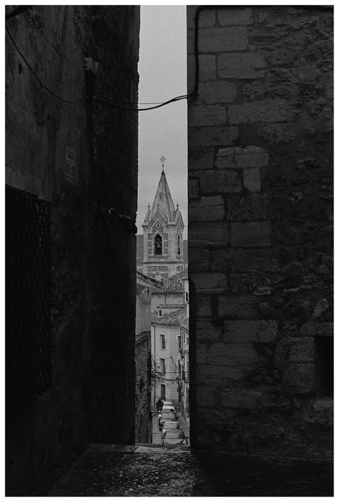 A narrow alleyway with dark stone walls frames a view of a church steeple and a street with parked cars.