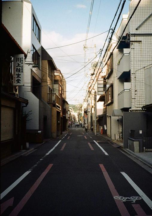 A narrow street in Japan is lined with buildings and overhead power lines.