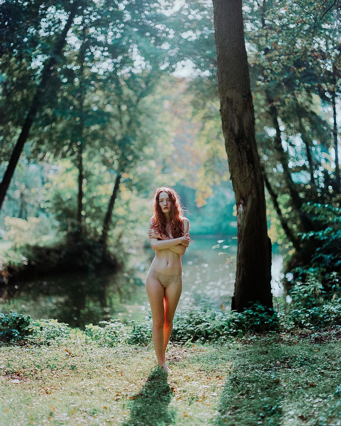 A woman with long red hair stands in a forest next to a body of water, with sunlight filtering through the trees.