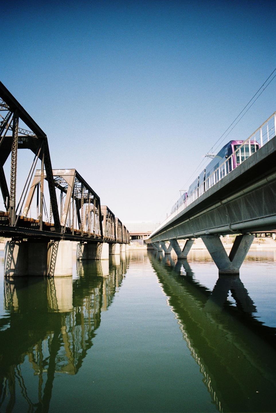A purple train crosses a bridge over a body of water next to a metal truss bridge.