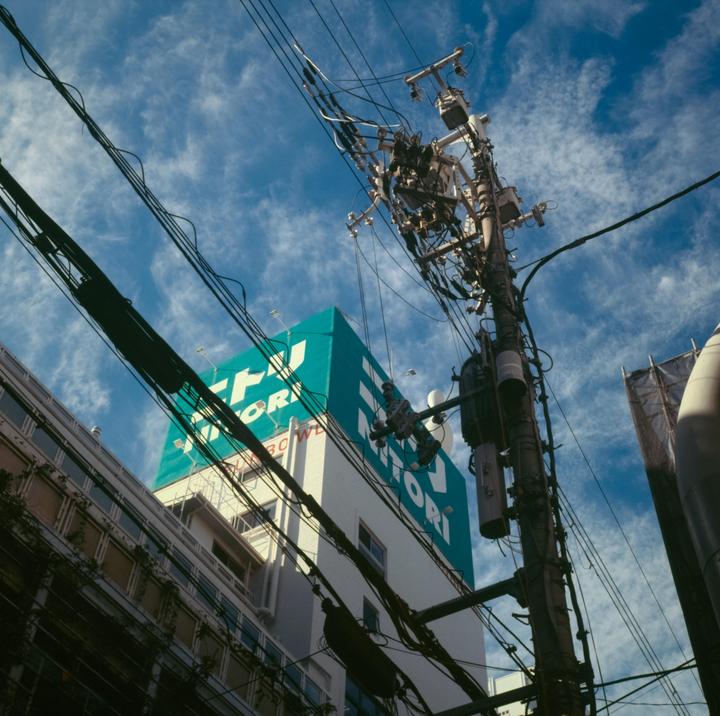 A low-angle view shows a utility pole with many wires against a blue sky, with a building and a large sign in the background.