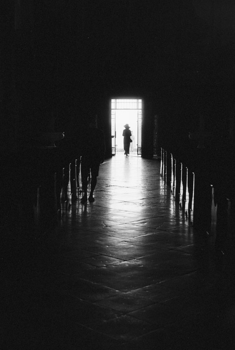 A woman in a hat stands in a bright doorway at the end of a dark hall with rows of benches.