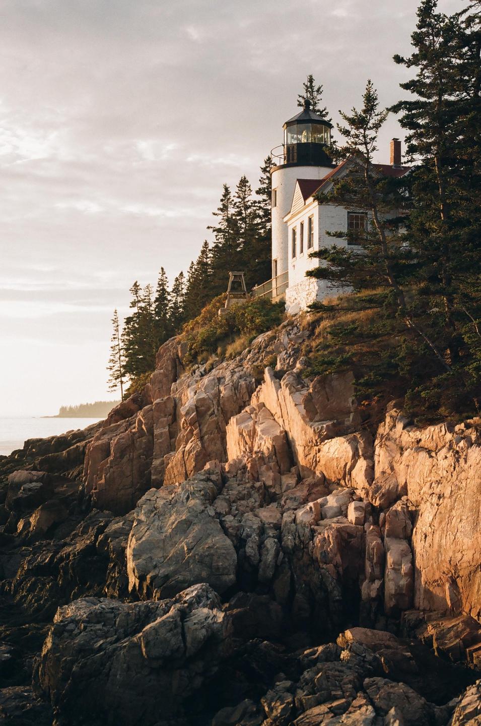 A white lighthouse and house stand on a rocky cliff overlooking the ocean at golden hour.