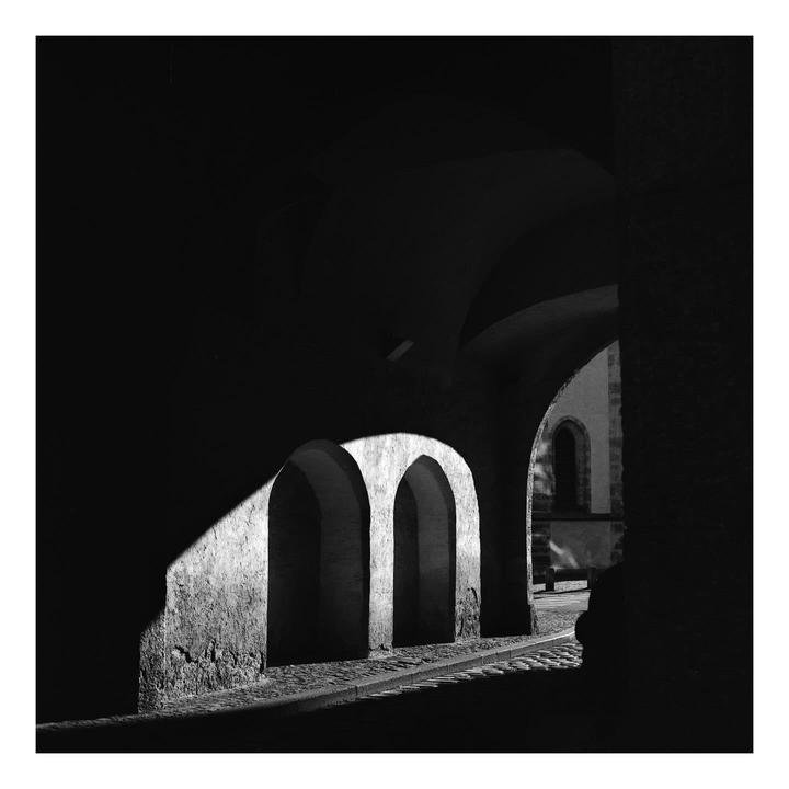 Sunlight illuminates a cobblestone street and three arches in a dark underpass.