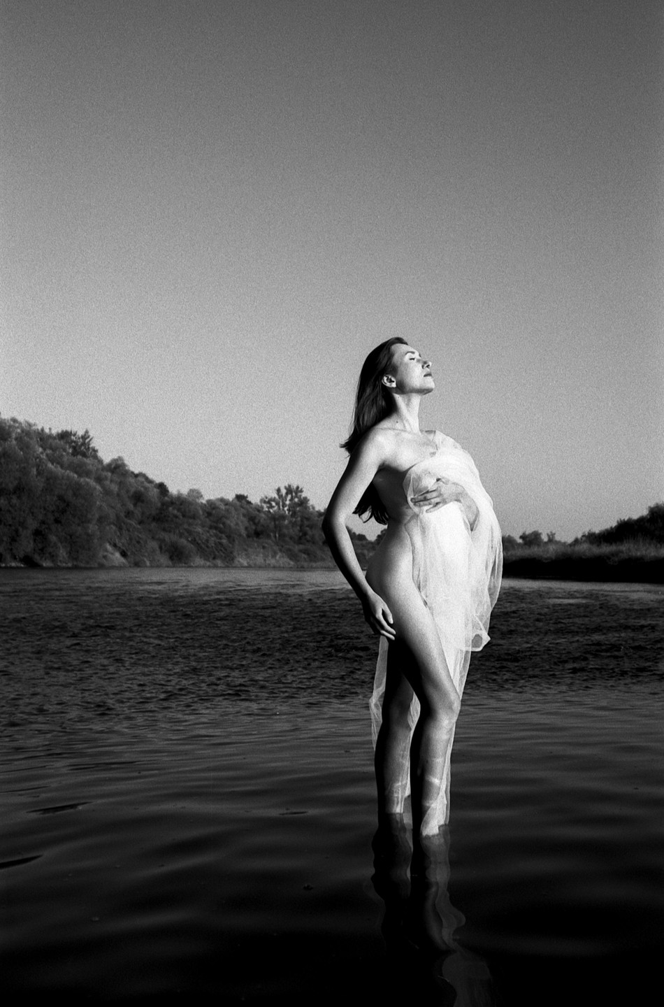 A woman stands in a river, partially draped in sheer fabric, with her head tilted back towards the sky.