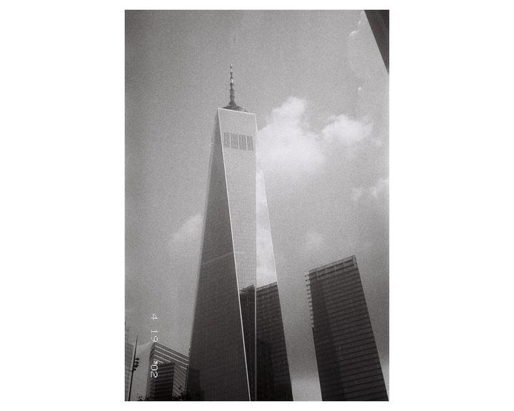 A low-angle view shows a tall, modern skyscraper with a spire against a cloudy sky, with other buildings visible in the background.