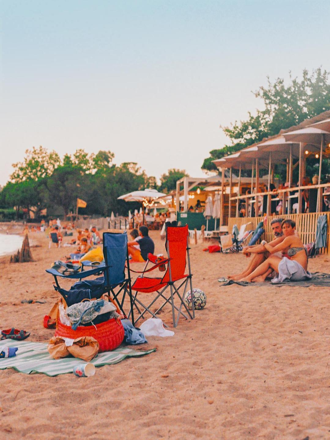 A couple sits on a sandy beach near a beach bar with other people and beach chairs.