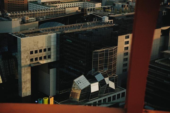 An aerial view shows a dense urban landscape with various buildings and structures, including a distinctive building with a geometric, fragmented roof design.
