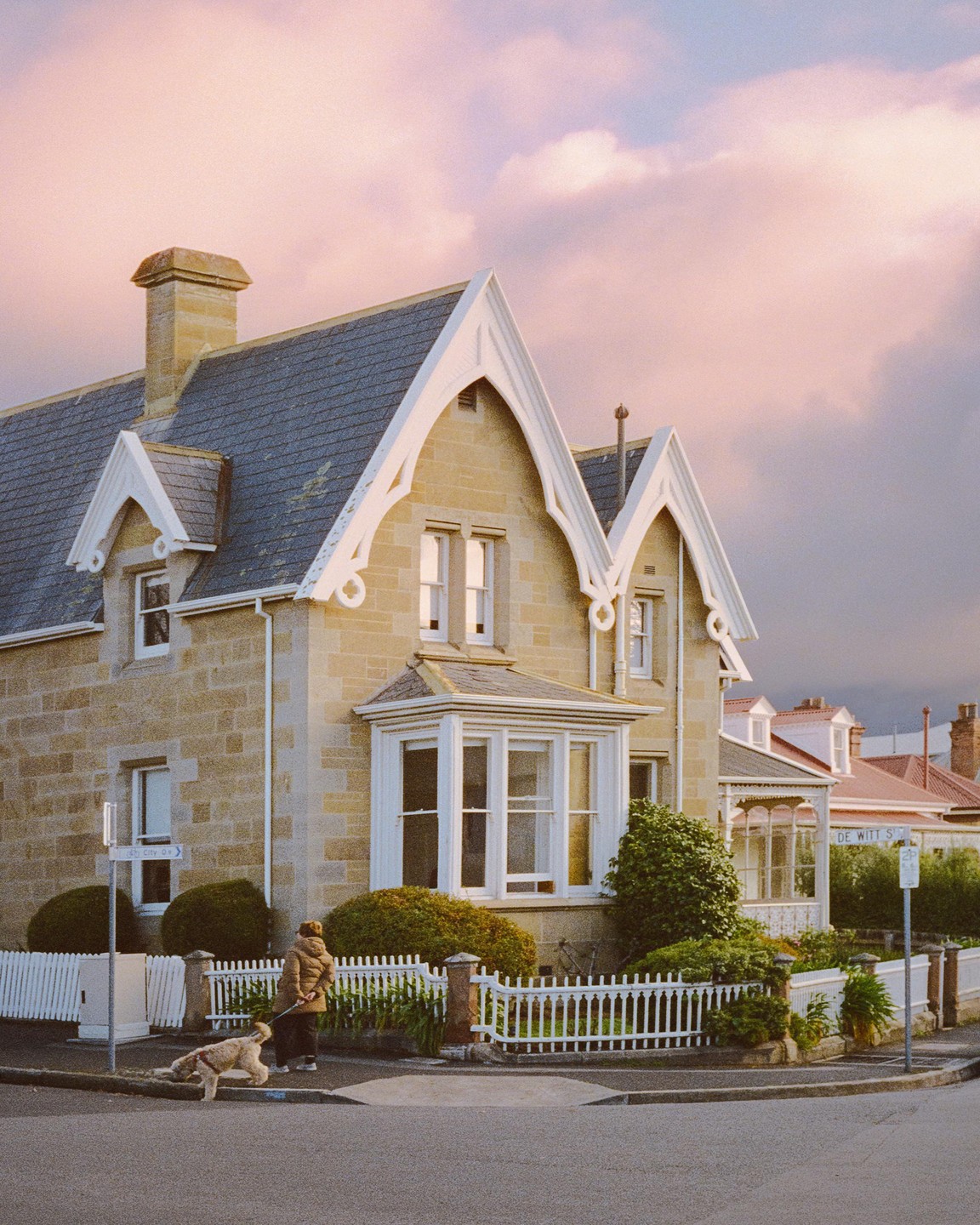 A person walks a dog past a large stone house with a white picket fence under a pink sky.