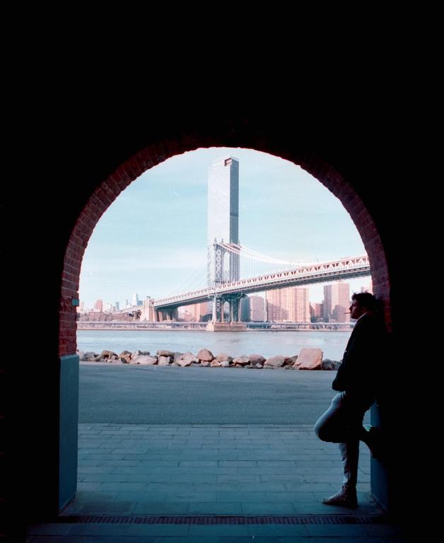 A man leans against a brick archway, looking out at a bridge and city skyline across a body of water.