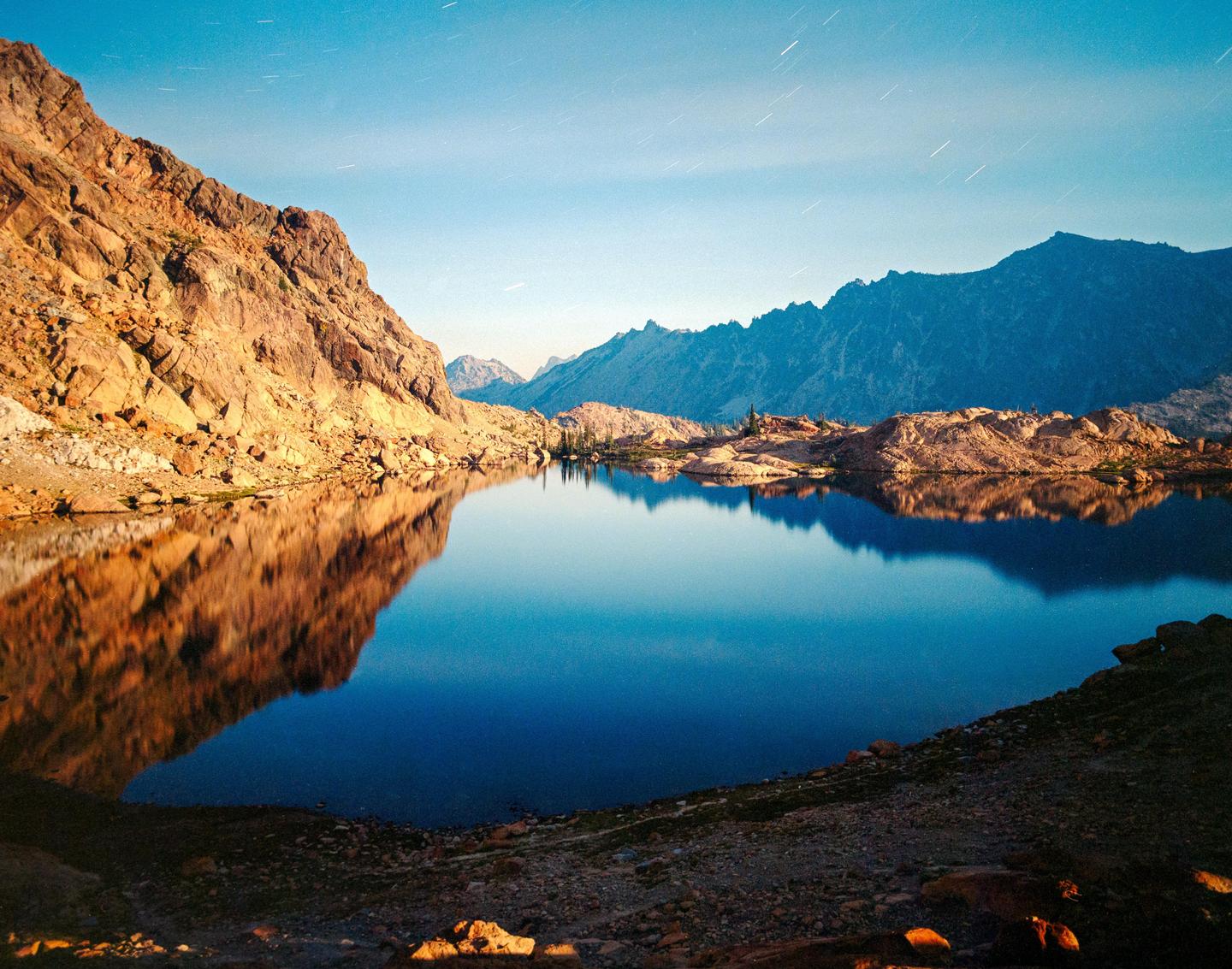 A long exposure shot of a lake at midnight shows star trails in the sky and mountains reflected in the water.