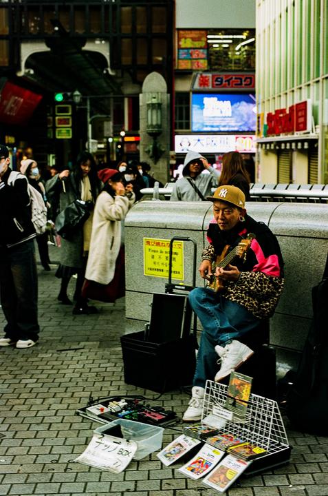 A man wearing a leopard print jacket and a baseball cap plays a ukulele on a city street.