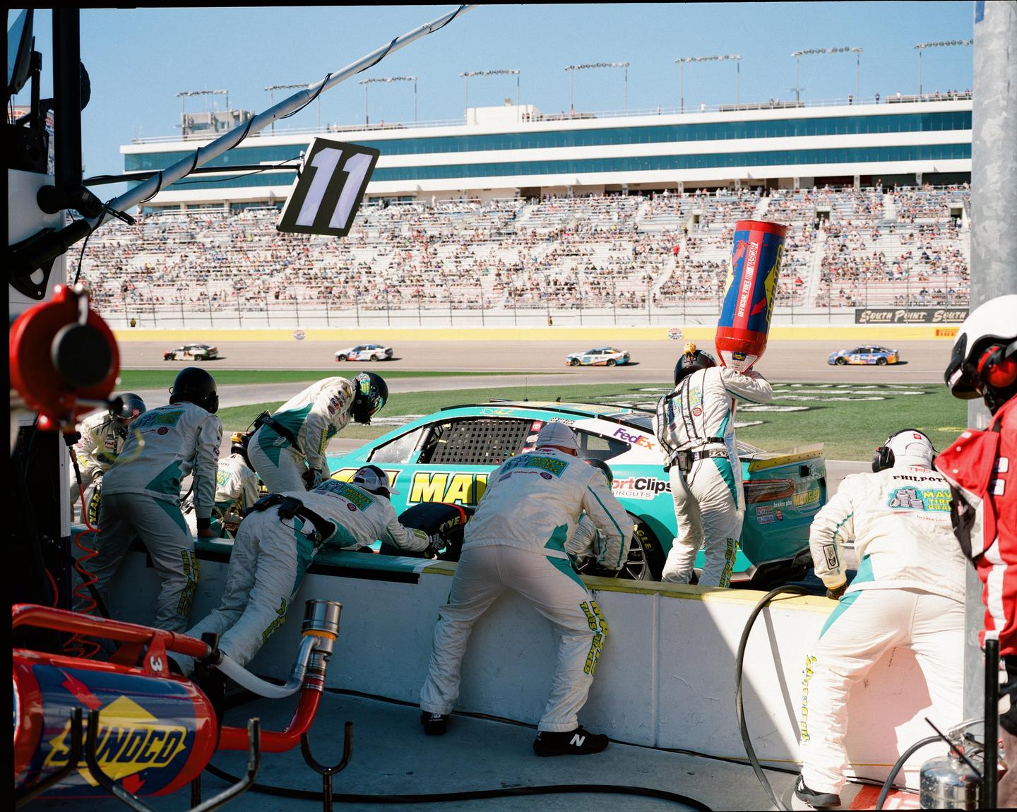 A pit crew services a race car on a track with a grandstand full of spectators in the background.