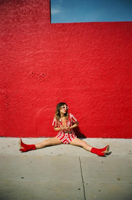 A woman in a red and white striped romper and red boots sits on the ground with her legs spread in front of a bright red wall.
