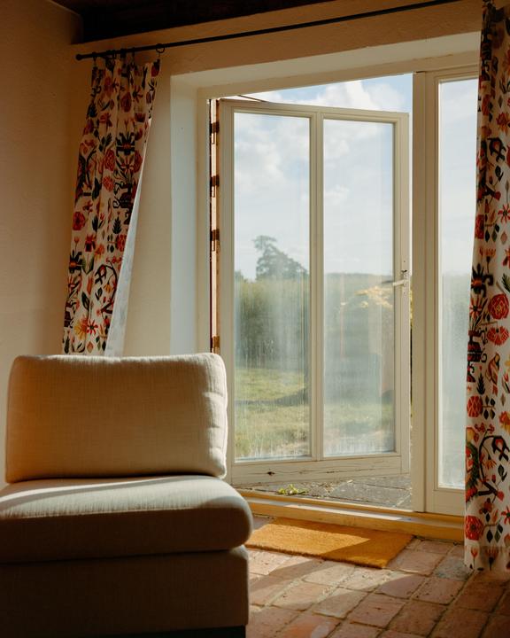 A beige armchair sits on a brick floor next to an open glass door with patterned curtains.
