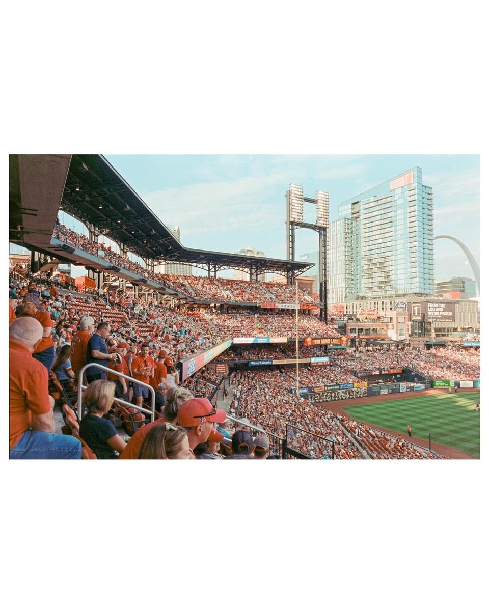 A baseball stadium is filled with spectators watching a game, with a city skyline visible in the background.
