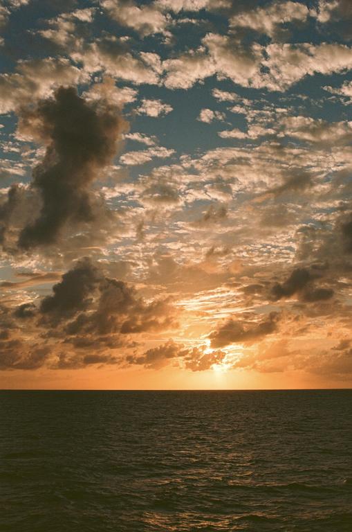 The sun sets over the ocean, casting an orange glow on the clouds and water.