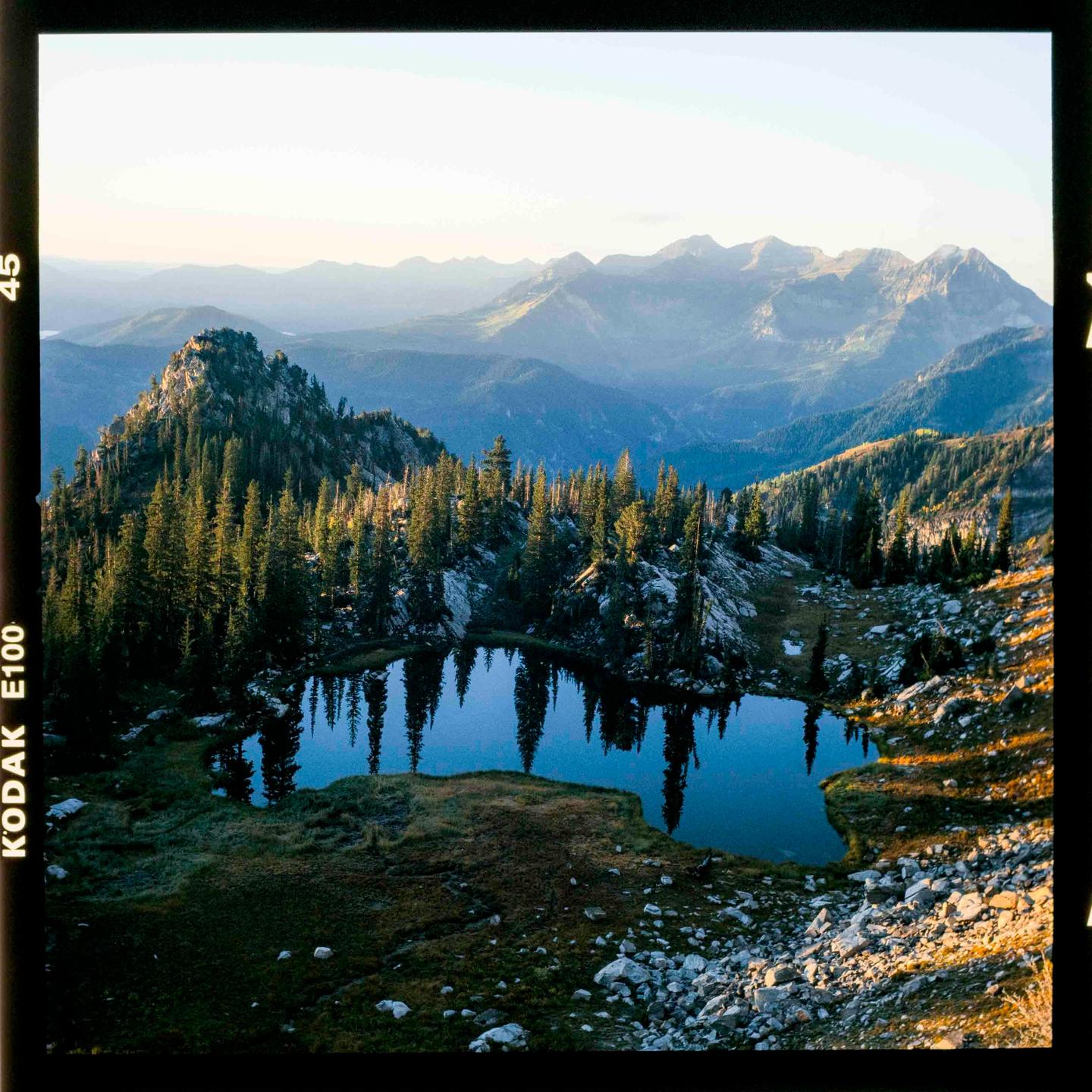 A mountain lake reflects pine trees under a clear sky with distant mountains.