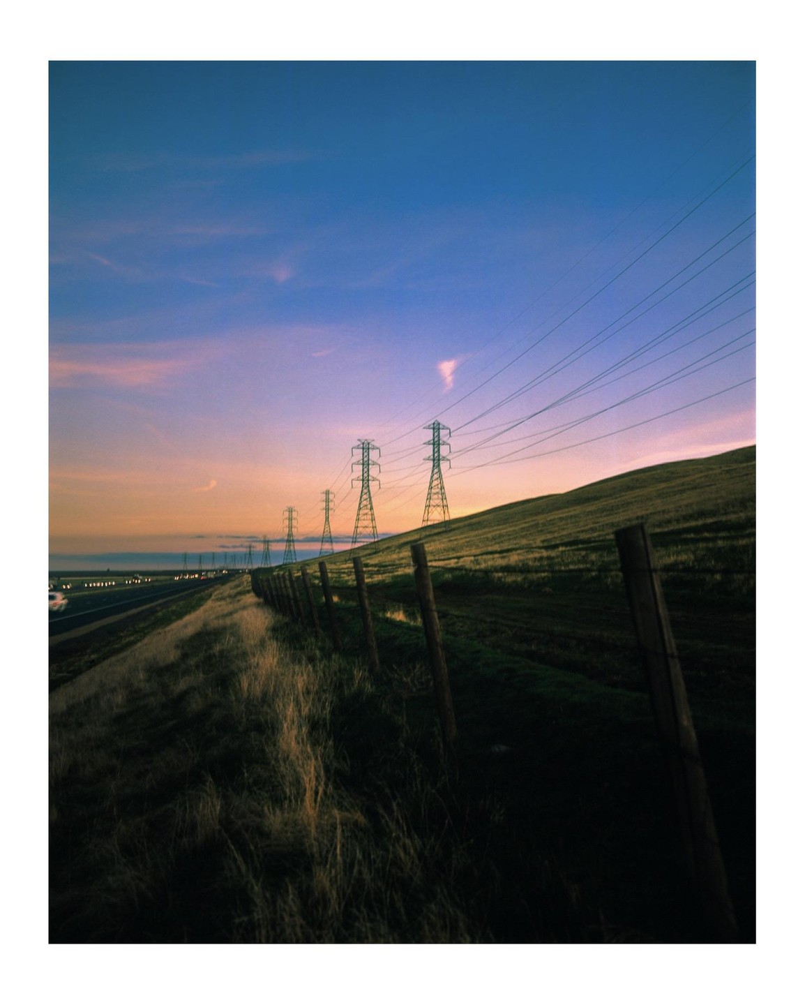 A row of power lines runs alongside a freeway and a grassy hill at dusk.
