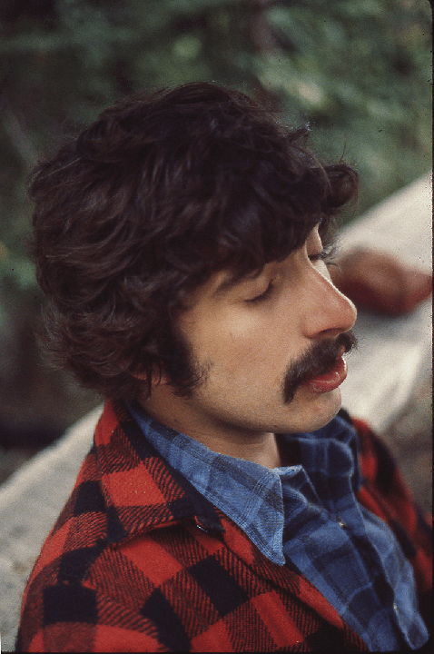 A man with a mustache and curly hair wears a red and black plaid jacket over a blue plaid shirt.
