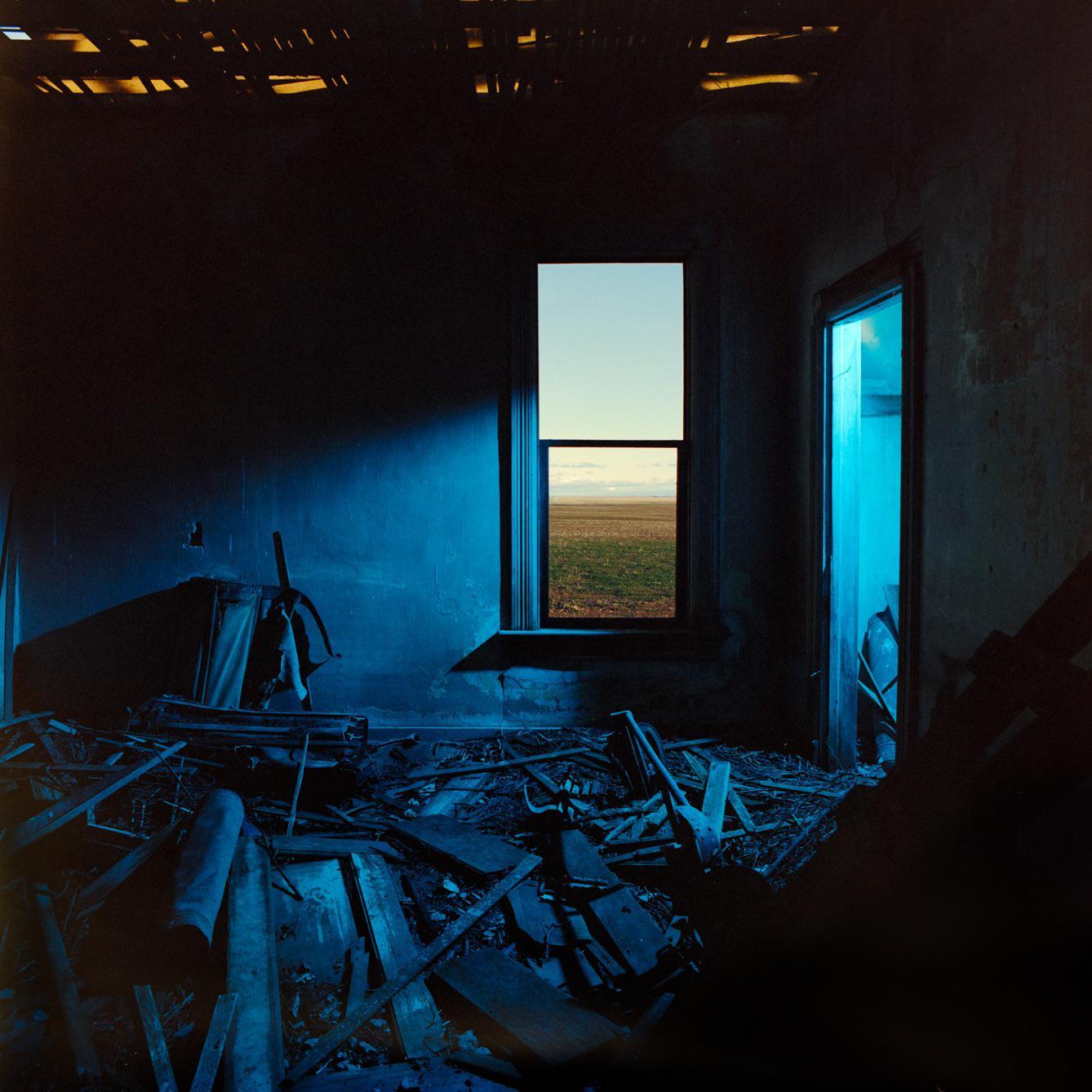 A dark, ruined room with blue light contains a window looking out onto a field.