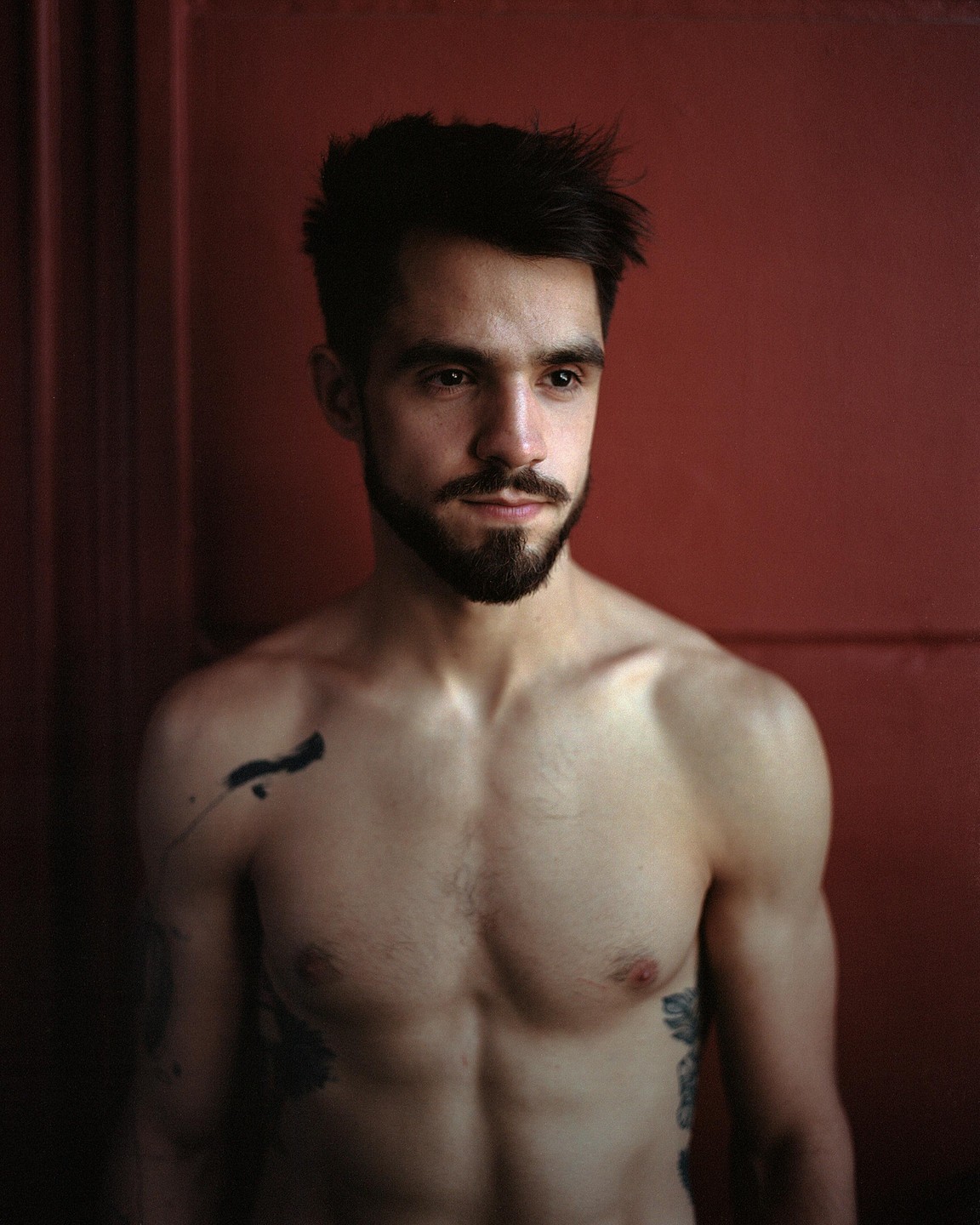A man with a beard and tattoos stands shirtless against a red wall.