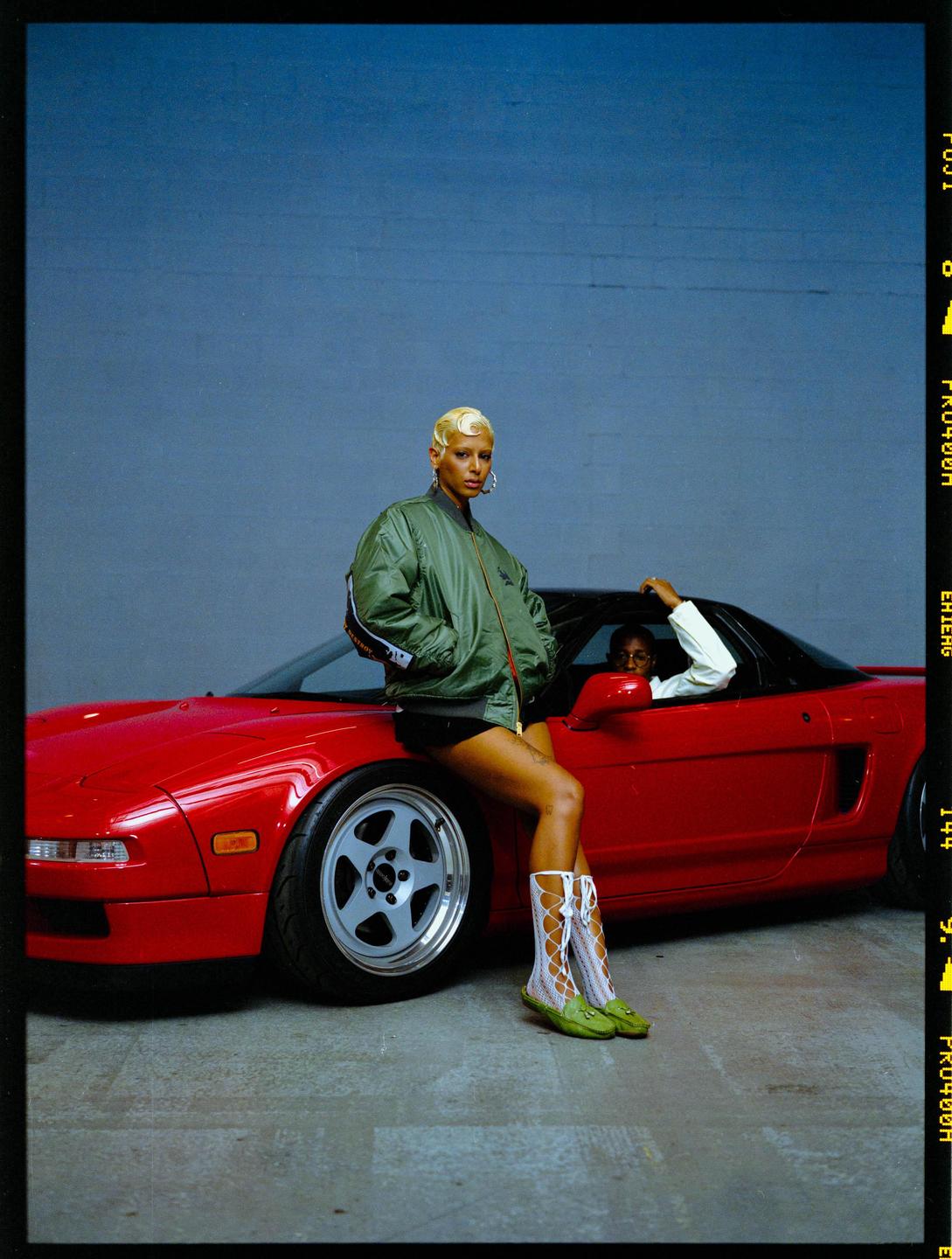 A woman with short blonde hair leans on a red sports car while a man sits in the driver's seat.