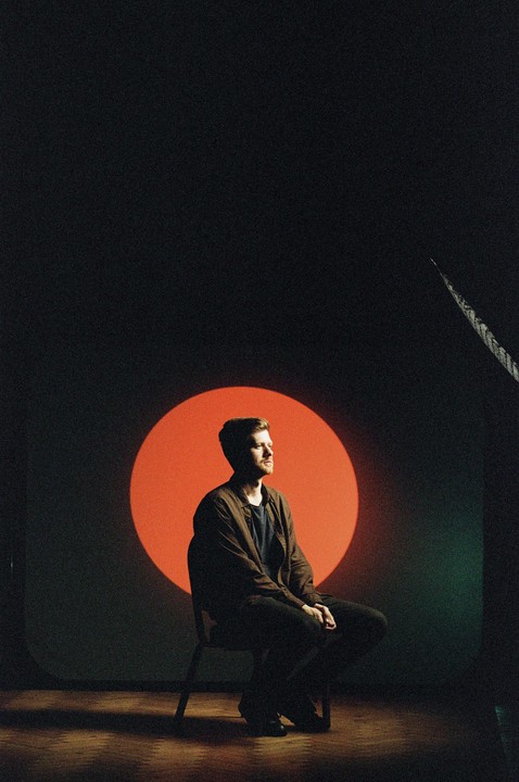 A man sits on a chair in front of a red circle projected onto a dark wall.