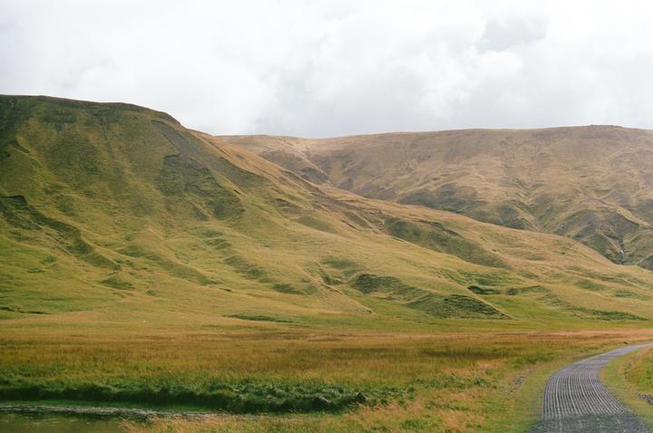 A gravel road curves through a grassy field with rolling hills under a cloudy sky.