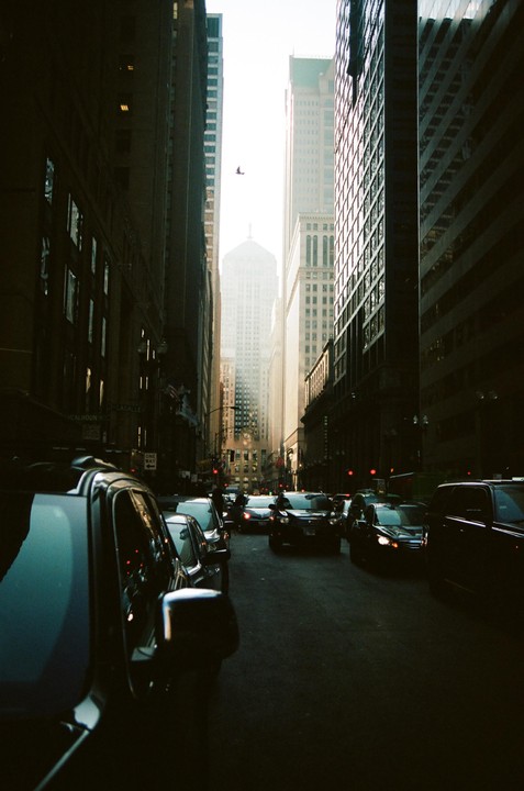 A street is filled with cars and tall buildings under a bright sky.