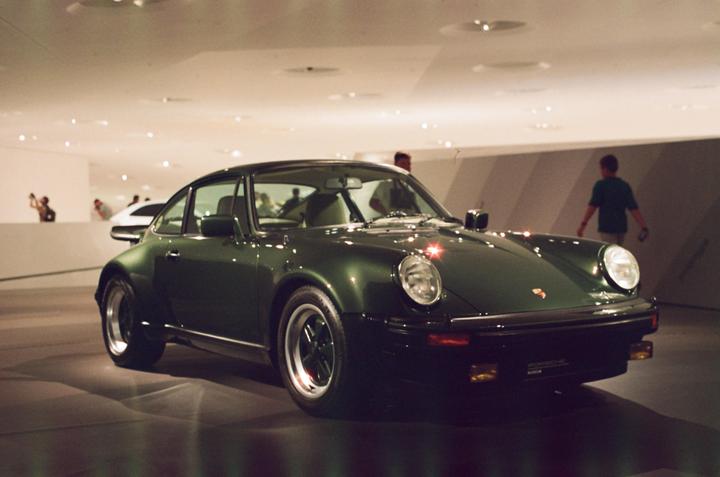 A dark green vintage car is parked in a brightly lit exhibition hall with people in the background.