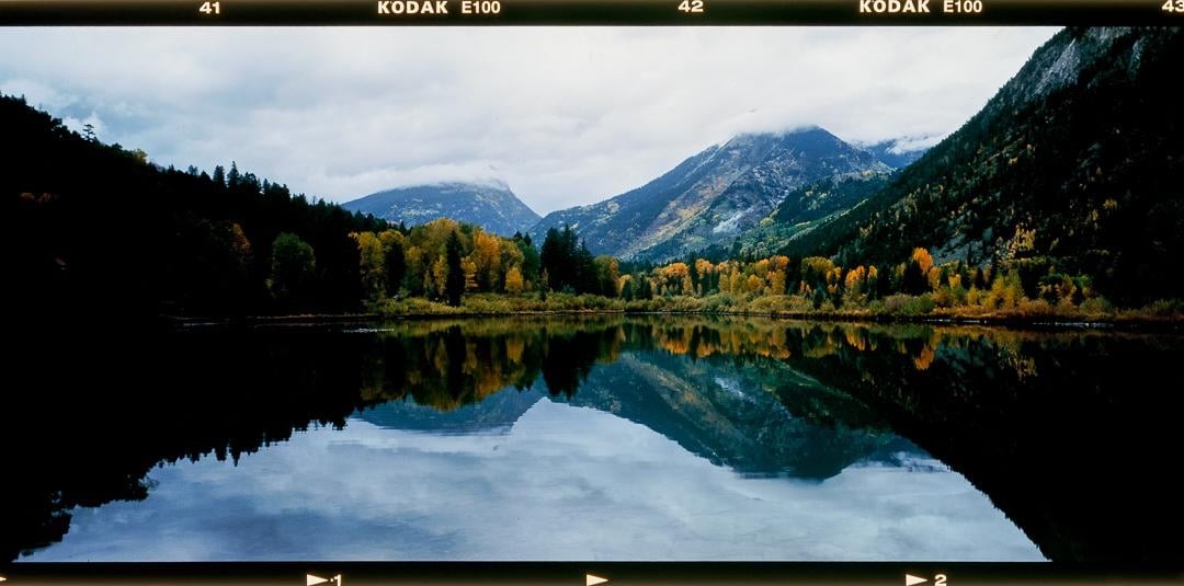 A panoramic landscape shows a lake reflecting mountains and autumn trees under a cloudy sky.
