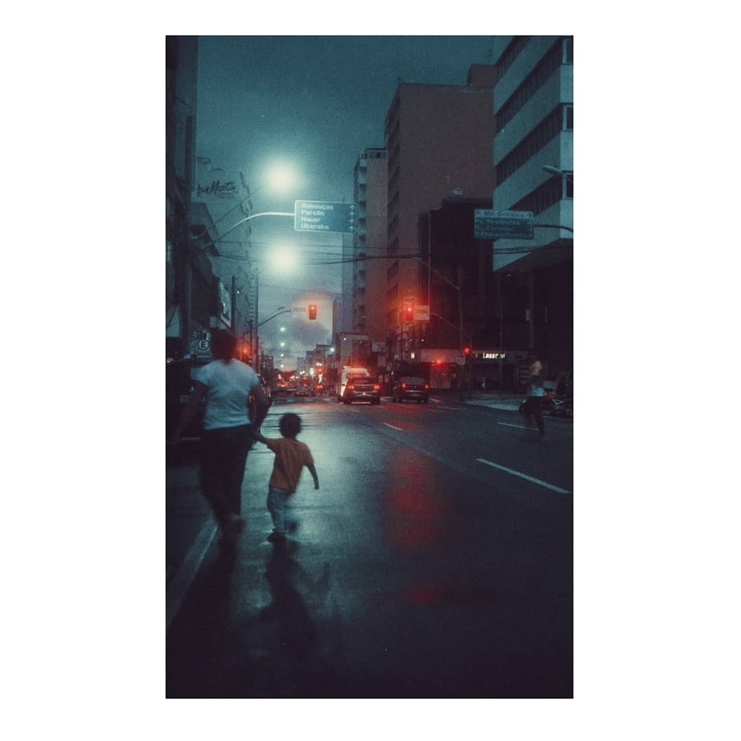 A woman and child walk hand-in-hand down a wet city street at night, with cars and buildings visible in the background.