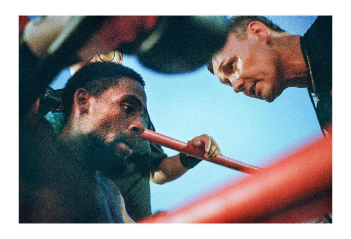 A man in a boxing ring looks down while another man leans over the ropes to speak to him.