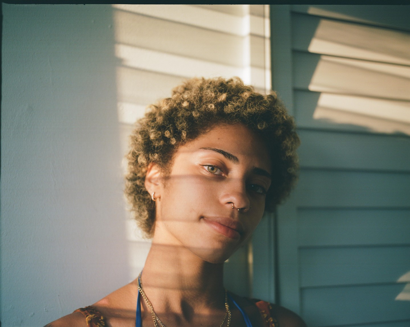 A woman with curly hair and a nose ring looks at the viewer, with shadows from blinds falling across her face.