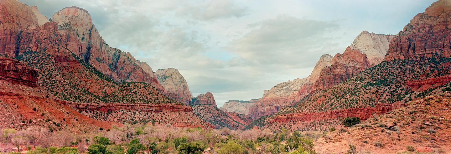 A panoramic landscape shows a valley with green trees and bushes surrounded by red and white mountains under a cloudy sky.